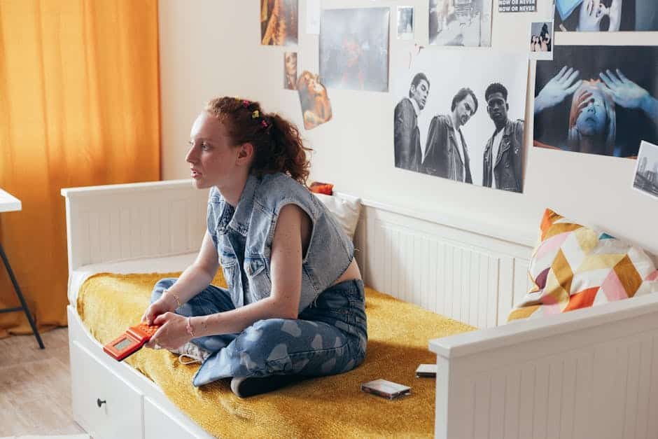 A woman in a denim vest sits on a bed holding a gamepad, surrounded by art posters.